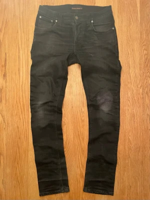 Nudie Jeans - Ett par svarta Slim fit jeans från Nudie Jeans i modell Grim Tim. De har tydliga slitningar och är både lagade i vecket och skrevet. De har ett minimalt hål i fickan samt hål utanför fickan (bild 4), genom hålet syns inte ens lår pga dess placering vid fickan. Förutom det finns inga defekter. Strl W29 L32.