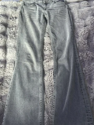 Grå bootcut jeans med slitningar - Säljer ett par grå bootcut jeans med snygga slitningar och klassisk femficksmodell. Jeansen har breda benslut och normal midja. Perfekta för dig som gillar retrovibbar och vill ha ett par bekväma jeans i mjukt denimtyg.
