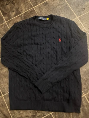 Mörkblå kabelstickad tröja Polo Ralph Lauren - Knappt använd, inte riktigt min stil. OBS DEN ÄR BLÅ, SER SVART UT PGA LJUS. Kabelstickad mörkblå tröja från Polo Ralph Lauren med rund halsringning och den klassiska röda logotypen broderad på bröstet. Tröjan har långa ärmar och ribbade muddar vid ärmslut och nederkant. Perfekt för dig som gillar tidlös och stilren design.