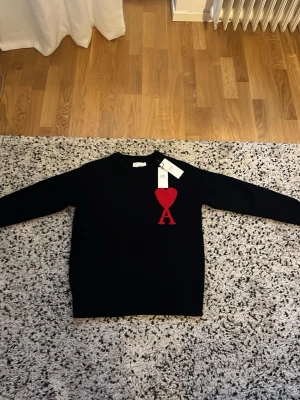 Svart stickad tröja från Ami Paris  - Snygg svart stickad tröja från Ami Paris med röd hjärtlogga och bokstaven A på bröstet. Tröjan har rund halsringning, långa ärmar och ribbade muddar vid ärmslut och nederkant. Perfekt för dig som gillar stilrena plagg med en twist. Men stoleken är M men sitter som en S. Alltså den är liten i storleken.