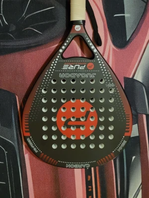 Padelracket JUGADOR Pure Carbon - Snyggt padelracket från JUGADOR, modell Pure, i svart och rött med vit logga. Racketen har droppform, många hål och grepp i ljust material. Tillverkat i kolfiber (carbon) för extra hållbarhet och prestanda. Perfekt för dig som vill ta spelet till nästa nivå.