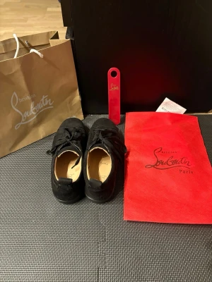 Svarta sneakers från Christian Louboutin - Stilrena svarta sneakers från Christian Louboutin i mocka med klassisk röd sula och svarta skosnören. Låg modell med rund tå och diskret branding på sulan. Perfekta för dig som vill ha en lyxig touch på din outfit.