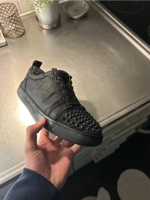Christian louboutins - Säljer ett par svarta sneakers från Christian Louboutin med coola svarta nitar över hela tån och klassiska svarta skosnören. Skorna har en låg profil och är tillverkade i läder med en snygg och edgy look. Kommer med kartong och skriv för snabb affär