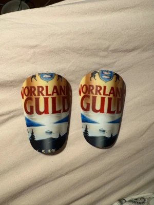 Norrlands Guld benskydd - Unika benskydd med Norrlands Guld-motiv. Framsidan har tryck i gult, blått och rött med landskapsmotiv och logga, baksidan är svart och vadderad för komfort. Perfekta för dig som vill sticka ut på planen! Benskydden är 8*5