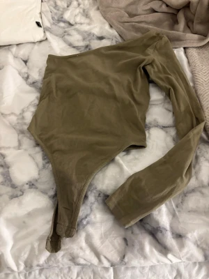 Khakigrön oneshoulder body från NA-KD - Säljer en khakigrön body från NA-KD i storlek XS. Modellen är oneshoulder med lång ärm och har knäppning i grenen. Materialet känns mjukt och stretchigt, perfekt för att styla med jeans eller kjol.