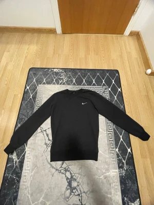 Svart Nike sweatshirt med logga - Svart sweatshirt från Nike med klassisk broderad vit swoosh på bröstet. Tröjan har rund hals, långa ärmar och ribbade muddar. Tillverkad i mjuk bomullsmix som passar perfekt till chill eller streetwear.