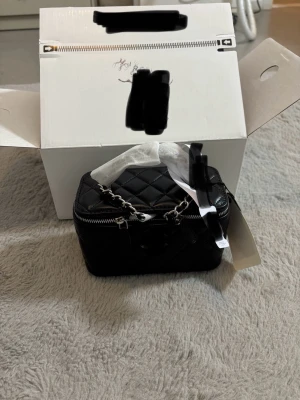 Svart quiltad handväska med kedja - Säljer en svart, rektangulär handväska med quiltat mönster och silverfärgad kedja som handtag. Väskan har dragkedja upptill och detaljer i skinn. Perfekt för dig som gillar stilrena accessoarer med lyxig känsla.