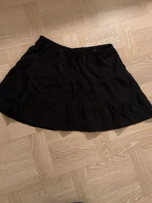 Svart volangkjol från H&M Divided - Kort svart kjol från H&M Divided med volangdetaljer och resår i midjan. Kjolen har en enkel och flowy passform som ger en avslappnad vibe. 