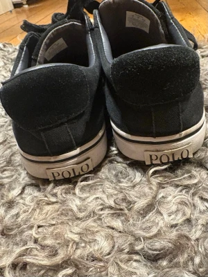Svarta sneakers från Polo Ralph Lauren - Säljer ett par svarta sneakers från Polo Ralph Lauren med klassisk vit sula och svart tyg. Skorna har snörning, rund tå och den ikoniska Polo-loggan broderad i guld på sidan. Perfekta till jeans eller shorts för en clean och stilren look. 