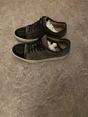 Grå sneakers från Lanvin med svart tå - Säljer ett par grå sneakers från Lanvin med svart lackad tå och svarta skosnören. Ovandelen är i mocka och sulan är vit. Snygg och stilren modell som passar till många outfits.
