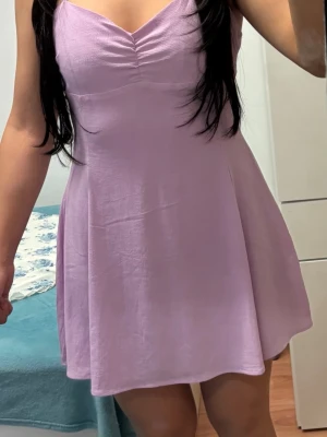 Ljuslila kort klänning med knyt - Supersöt ljuslila kort klänning med tunna axelband som knyts i nacken. Klänningen har en lätt v-ringning med rynkdetalj framtill och ett flowy fall. Perfekt för varma dagar och ger en fräsch vibe.