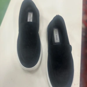 Svarta slip-on sneakers från Steve madden  - Svarta slip-on sneakers från Steve Madden med vit platt sula och rund tå. Ovandelen är i mocka och har en stilren design utan snörning. Diskret metallknapp bak på hälen och tydlig logga på sulan. Perfekt för dig som gillar enkel och snygg streetstil.