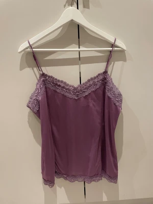 Lila linnetopp med spetsdetaljer - Säljer en lila linnetopp med tunna axelband och vacker spets längs urringning och nederkant. Toppen har en mjuk, glansig yta och avslappnad passform. Perfekt att styla med jeans eller kjol för en trendig look.