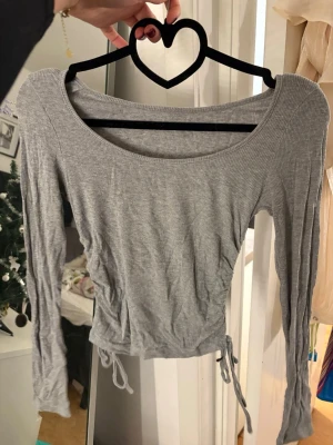 Grå ribbad långärmad topp - Säljer en grå ribbad långärmad topp med snörning i sidorna. Toppen har en rundad, bred urringning och är croppad i modellen. Perfekt att styla med höga jeans eller kjol för en trendig look. Den är från SHEIN och är i storlek S.