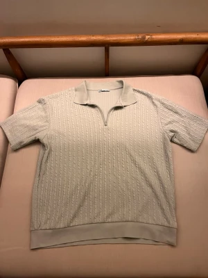 Beige kabelstickad polotopp från Zara - Säljer en beige kabelstickad polotopp från Zara i storlek M. Toppen har korta ärmar, krage och en half zip-dragkedja framtill. Materialet är mjukt och strukturerat med ribbade detaljer längst ner. 