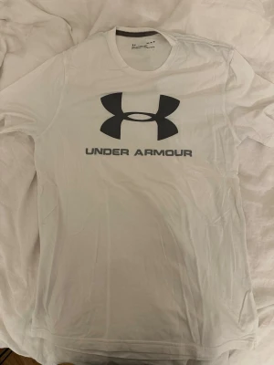 Vit Under Armour t-shirt loose fit - Säljer en vit t-shirt från Under Armour med stor svart logga och text på bröstet. Modellen är loose fit och tillverkad i ett lätt och funktionellt material, perfekt för träning eller chill. Klassisk rund hals och korta ärmar.