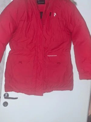 Röd Peak Performance parkajacka L - Peak Performance parkajacka i rött med huva och fuskpäls. Jackan har flera fickor, dragkedja och logga på bröstet. Perfekt för kalla dagar och har en klassisk, lite längre modell. Snygg och funktionell för vintern!