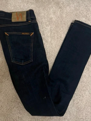 Mörkblå  Nudie Jeans - Säljer ett par mörkblå jeans från Nudie Jeans med klassiska orange sömmar och läderpatch bak. Jeansen har smal passform och raka ben, tillverkade i slitstarkt jeansmaterial. Perfekta för dig som gillar en clean och tidlös look.