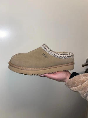 Beige UGG slip-on mockasiner - Säljer ett par beige slip-on skor från UGG med mjuk päls på insidan och mocka på utsidan. Skorna har en rund tå, platt sula och dekorativt mönster runt öppningen. Perfekta för att hålla fötterna varma och sköna under kyliga dagar.