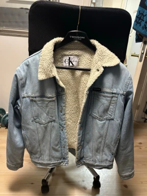 Calvin Klein ljusblå jeansjacka med teddyfoder - Säljer en fett snygg ljusblå jeansjacka från Calvin Klein Jeans med mjukt teddyfoder i offwhite. Jackan har klassisk krage, två bröstfickor med knappar och knappstängning framtill. Perfekt för dig som vill ha en chill och varm look.