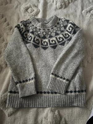 Grå stickad tröja med mönster - Säljer en grå stickad tröja med svart och vitt nordiskt mönster runt halsen och detaljer vid ärmslut och nederkant. Tröjan har rund hals och långa ärmar, perfekt för kalla dagar. Supermysig och klassisk stil!