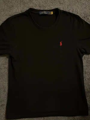 Svart t-shirt från Polo Ralph Lauren - Svart t-shirt från Polo Ralph Lauren med klassisk röd broderad logga på bröstet. T-shirten har rund halsringning och korta ärmar. Tillverkad i mjuk bomull för en skön känsla och clean look.