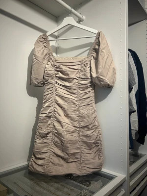 Rynkad miniklänning med puffärm NA-KD - Superfin rynkad miniklänning från NA-KD i beige med puffärmar och fyrkantig urringning. Klänningen har en figurnära passform och snygga rynkdetaljer framtill och baktill. Perfekt för dig som vill sticka ut med en trendig och stilren look. Använd 1 gång.