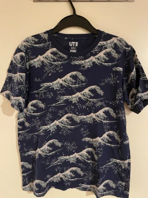 Blå Hokusai t-shirt från Uniqlo - Säljer en blå t-shirt från Uniqlo UT med Hokusai Blue-tryck. T-shirten har korta ärmar och är täckt av ett vågigt konstmotiv inspirerat av japansk konst. Tillverkad i mjuk bomull och har en avslappnad passform. Perfekt för dig som gillar unika prints.
