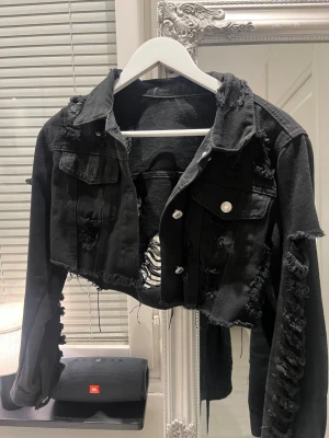 Svart croppad jeansjacka med slitningar - Säljer en svart croppad jeansjacka med råa kanter och tydliga slitningar över hela jackan. Jackan har silvriga knappar framtill, bröstfickor och lång ärm med slitna detaljer längs ärmarna. Perfekt för dig som gillar en edgy och trendig stil.