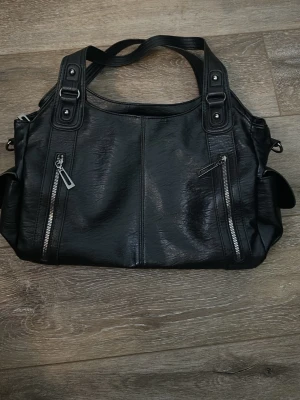 Svart axelväska med dragkedjor - Snygg svart axelväska i skinnimitation med flera dragkedjefickor både fram och bak. Väskan har breda axelband och 2 rymliga innerfack med extra fickor. Perfekt för dig som vill ha plats för allt du behöver under dagen.