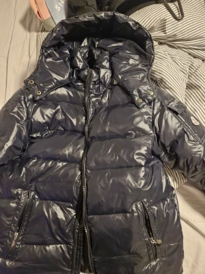 Mörkblå Moncler pufferjacka med huva - Säljer en svarr pufferjacka från Moncler med glansig finish, huva och dragkedja framtill. Jackan har praktiska fickor med dragkedja och en ficka med knapp på ärmen där Moncler-loggan sitter. Insidan har ett coolt serietidningsmotiv. Vill bara bli av med den pris kan diskuteras 