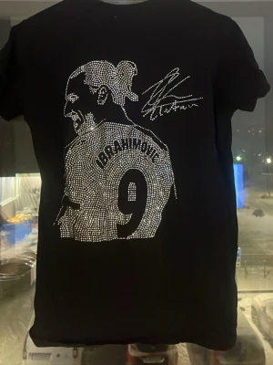 Svart t-shirt med strassmotiv Ibrahimovic - Svart t-shirt med korta ärmar och ett stort motiv av Ibrahimovic och nummer 9 på ryggen, gjort i glittrande strass. Snygg signaturdetalj upptill. Perfekt för dig som vill sticka ut och visa din kärlek för fotboll och ikoniska spelare.