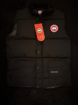 Svart dunväst från Canada Goose - Säljer en svart dunväst från Canada Goose med klassisk logga på bröstet och diskret broderad text på fickan. Västen har tryckknappar, dragkedja och två stora fickor framtill. Perfekt för kalla dagar och riktigt snygg till lager-på-lager.
