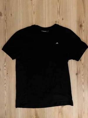 J.Lindeberg cotton blend t shirt - strl.S - En stilren svart t-shirt från J.Lindeberg med liten vit logga på bröstet. Klassisk passform och rund halsringning, perfekt till jeans eller shorts. Tillverkad i mjuk bomull för skön känsla hela dagen.
