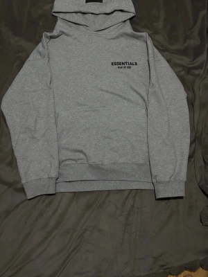 Grå Essentials hoodie - Den här hoodien är ljusgrå och har en stilren, minimalistisk design. På ryggen sitter den svarta texten, ESSENTIALS FEAR OF GOD tryckt i ett tydligt och modernt typsnitt. Materialet är mjukt och tjockt, vilket ger ett bekvämt och värmande intryck. Den är perfekt för vardagen om man inte vill ha något för stiligt men också att det ska vara snyggt för den vanliga vardage