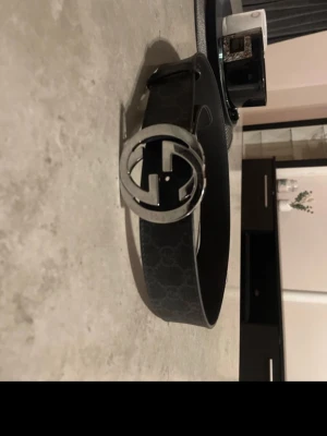 Svart Gucci skärp med GG-spänne - Säljer ett svart äkta skärp från Gucci med det klassiska GG-mönstret och en stor, rund metallspänne med GG-logga. Skärpet är gjort i skinn och har präglad logga på insidan. Perfekt accessoar för att lyfta vilken outfit som helst. Vissa skavanker finns (se på sista bilderna) Mått: 95cm