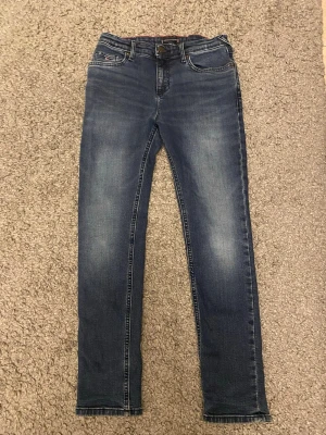 Blå skinny jeans Tommy Hilfiger - Säljer ett par klassiska blå skinny jeans från Tommy Hilfiger i storlek 152. Jeansen har fem fickor, snygga slitningar och ikoniska Hilfiger-detaljer på bakfickan och knappen. Perfekt passform med normal midja och stretchigt jeansmaterial.