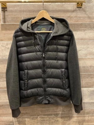 Polo Ralph Lauren cardigan  - Säljer en svart pufferjacka från Polo Ralph Lauren med rutiga ärmar och huva. Jackan har dragkedja framtill, två fickor med knapp och quiltad kropp. Materialmix med polyester och rutigt tyg på ärmar och huva ger en unik look. Perfekt för dig som vill ha något stilrent men ändå annorlunda.