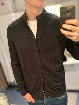 Mörkgrå kofta med knappar  - Snygg mörkbrun kofta med lång ärm och raka muddar. Koftan har en stilren ståkrage och knäpps med svarta knappar hela vägen framtill. Tillverkad i mjukt material som känns skönt mot huden. Perfekt att slänga över en t-shirt för en clean look.