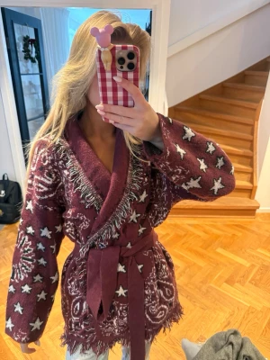 Vinröd kofta med stjärnor och bälte - Säljer en vinröd kofta med vita stjärnor och paisleymönster. Koftan har fransade kanter, knyts med bälte i midjan och har bred krage. Materialet är stickat och den har en loose passform med långa ärmar. Perfekt för höst och vinter!