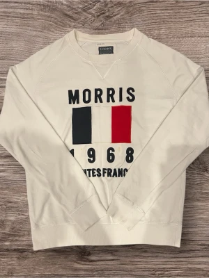 Vit sweatshirt från Morris, M - Vit sweatshirt från Morris med tryck framtill i blått, vitt och rött samt texten 'Morris 1968 Nantes France'. Tröjan har rund hals, långa ärmar och ribbade muddar. Perfekt för en chill och stilren look.
