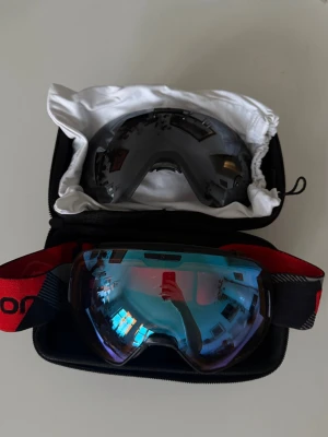 Salomon skidglasögon med extra lins - Säljer ett par Salomon skidglasögon med spegelblå lins och svart-rött band med logga. Medföljer extra mörk lins och skyddande case. Sitter bekvämt och har bred sikt, perfekt för dig som vill ha stilrena goggles i backen.