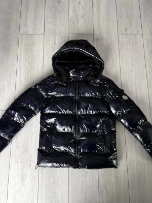 Mörkblå pufferjacka från Moncler - Säljer en glansig mörkblå pufferjacka från Moncler med huva och dragkedja framtill. Jackan har två fickor med dragkedja och en patch med Moncler-logga på ärmen. Perfekt för kalla dagar och har en riktigt snygg streetstil.
