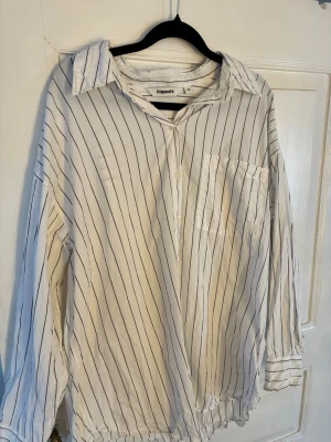 Vit randig skjorta från Kappahl - Vit skjorta från Kappahl i storlek M med svarta vertikala ränder. Skjortan har klassisk krage, bröstficka och knäppning framtill. Långa ärmar med manschett och knapp. Tillverkad i mjuk bomull, perfekt för en chill och stilren look.