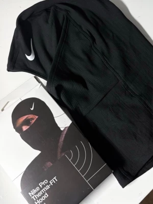 Nike Pro Therma-FIT svart balaclava - Nike Pro Therma-FIT balaclava i svart med vit Nike-logga. Tillverkad i ett syntetmaterial som håller värmen och är perfekt för träning utomhus, speciellt fotboll. Sitter tight mot huvudet och täcker både hals och ansikte.