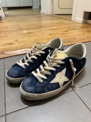 Golden Goose Super-Star blå sneakers - Golden Goose Super-Star sneakers i blå mocka med vit stjärndetalj på sidan och silvriga hälpartier. Skorna har vita snören med Golden Goose-tryck och en klassisk låg siluett. Insidan är vit och sulan är platt och vit. Perfekta för dig som gillar streetstyle och premiumkänsla.
