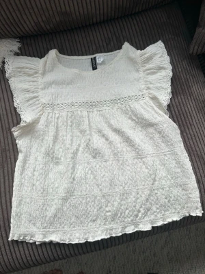 Vit blus med spets från H&M Divided - Supersöt vit blus från H&M Divided i storlek M. Blusen har korta volangärmar, spetsdetalj över bröstet och är gjord i ett lätt, krinklat bomullstyg. Perfekt till sommaren och enkel att styla med jeans eller kjol. Nästan aldrig använd och liten i storleken!