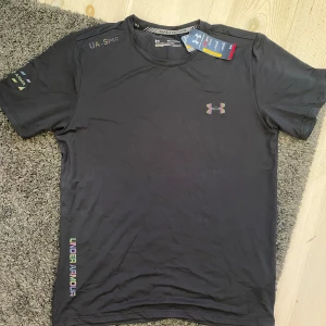 Svart Under Armour t-shirt - Snygg svart t-shirt från Under Armour med färgglada loggor på bröstet och vid nederkanten. T-shirten har rund hals och korta ärmar, perfekt för träning eller chill. Materialet är lätt och andas, vilket gör den skön att ha på sig under aktiva dagar.