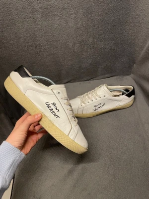Saint Laurent skor - Säljer ett par klassiska vita sneakers från Saint Laurent med svart detalj på hälen och handskriven logga på sidan i storlek 40 (passar även 41). Skorna är Tillverkade i skin. Nypris ligger på runt 8000. Skorna har tecken på andvändning                                                            Skriv vid frågor, priset är inte hugget i sten🤝