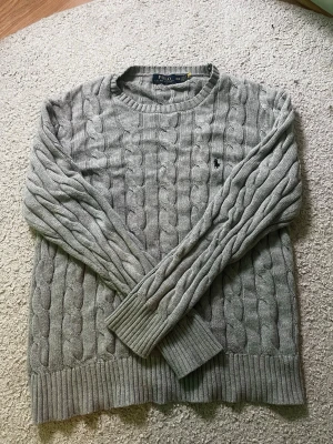  Polo Ralph Lauren - Säljer en grå kabelstickad tröja från Polo Ralph Lauren i storlek M. Tröjan har rund hals, ribbade muddar och det klassiska broderade Polo-logot på bröstet. Perfekt för dig som gillar stilrena och tidlösa plagg med snygg struktur.